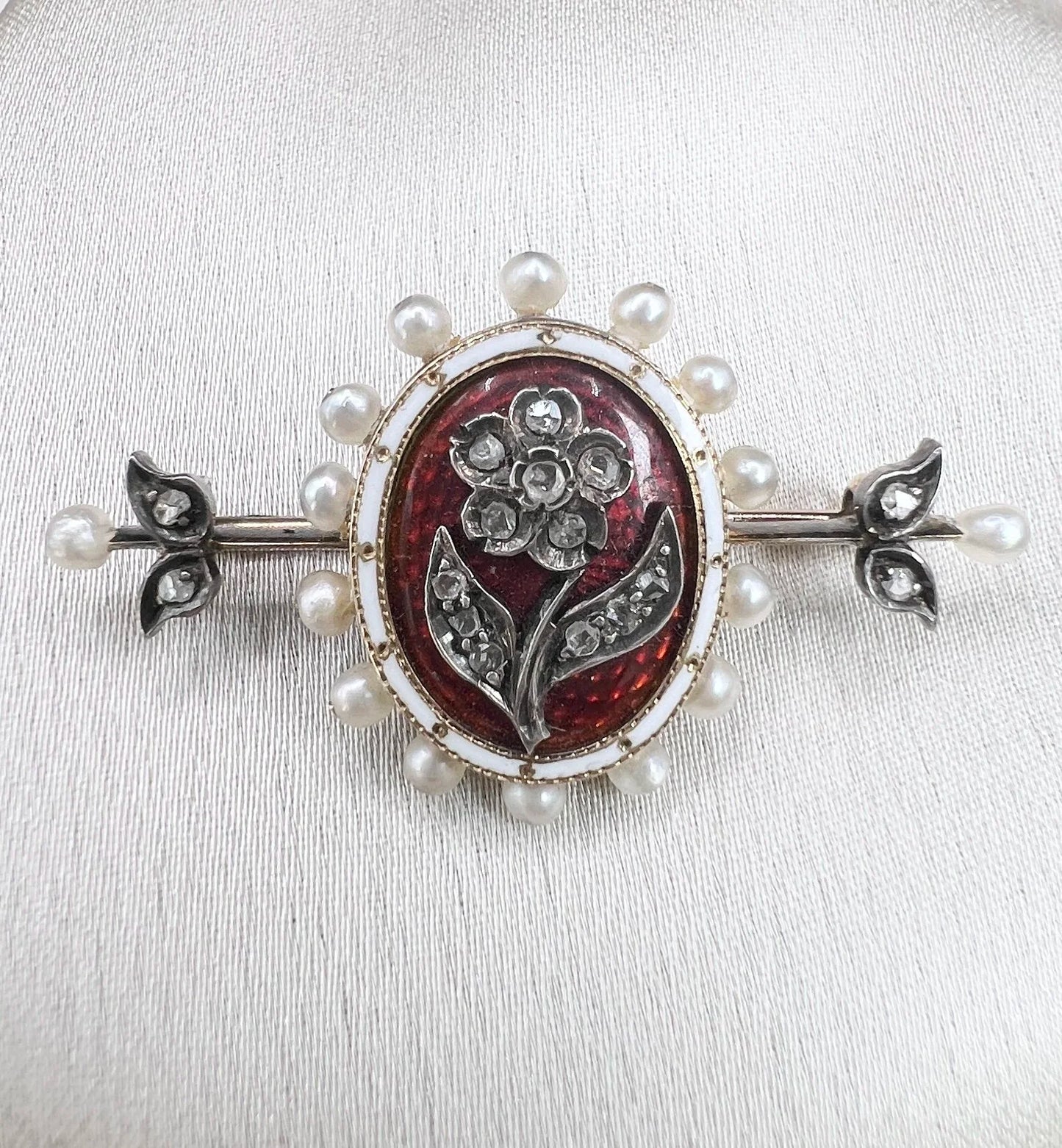 A yellow gold bar brooch with a red enamel centre surrounded by pearls, with a diamond flower motif and diamond leaves at either end