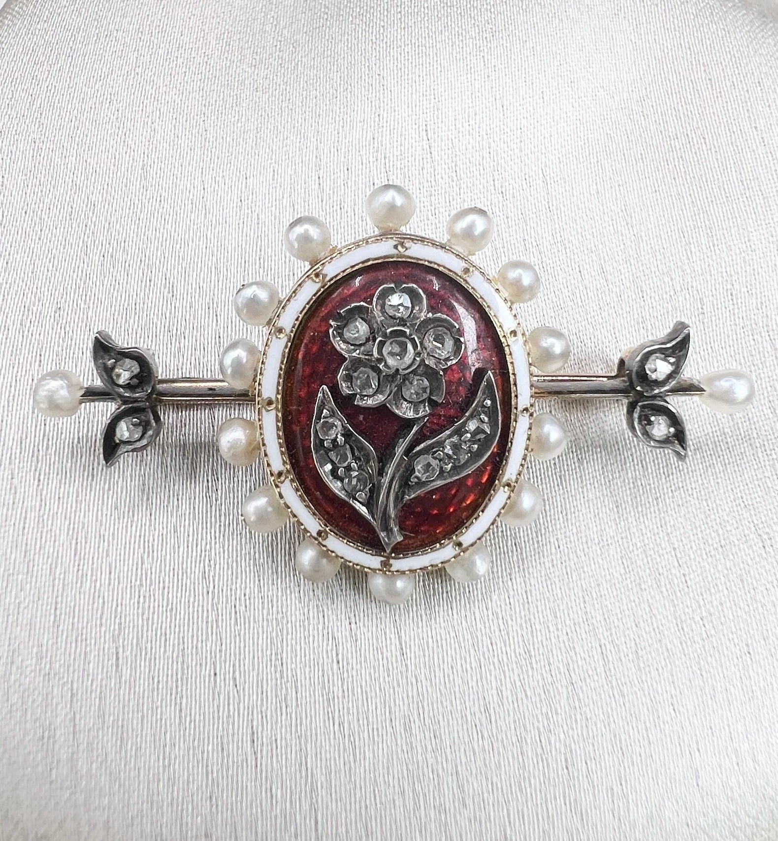 A yellow gold bar brooch with a red enamel centre surrounded by pearls, with a diamond flower motif and diamond leaves at either end