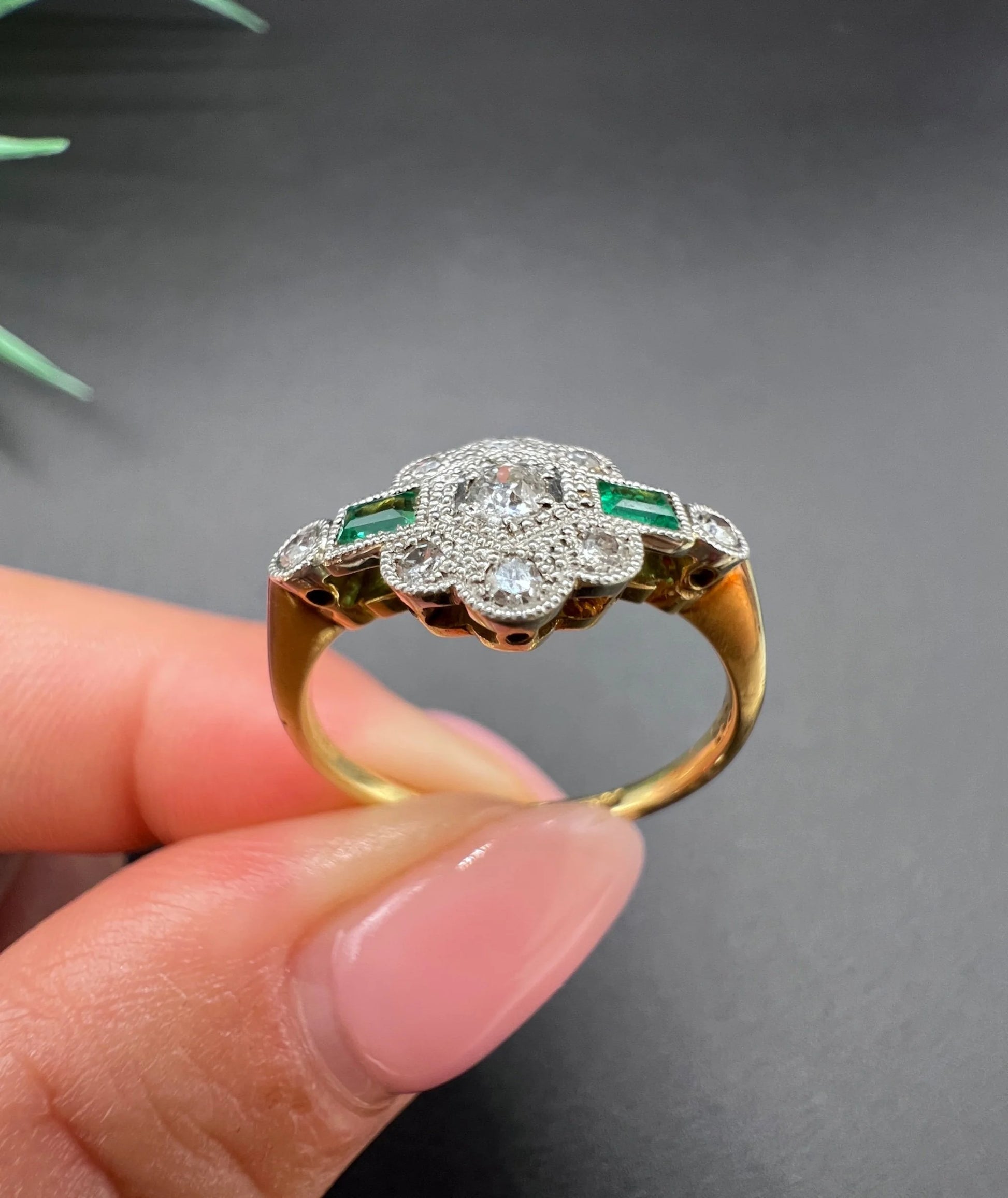 An emerald and diamond floral cluster ring in yellow gold and platinum, held in a hand