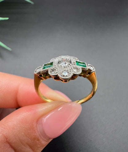 An emerald and diamond floral cluster ring in yellow gold and platinum, held in a hand