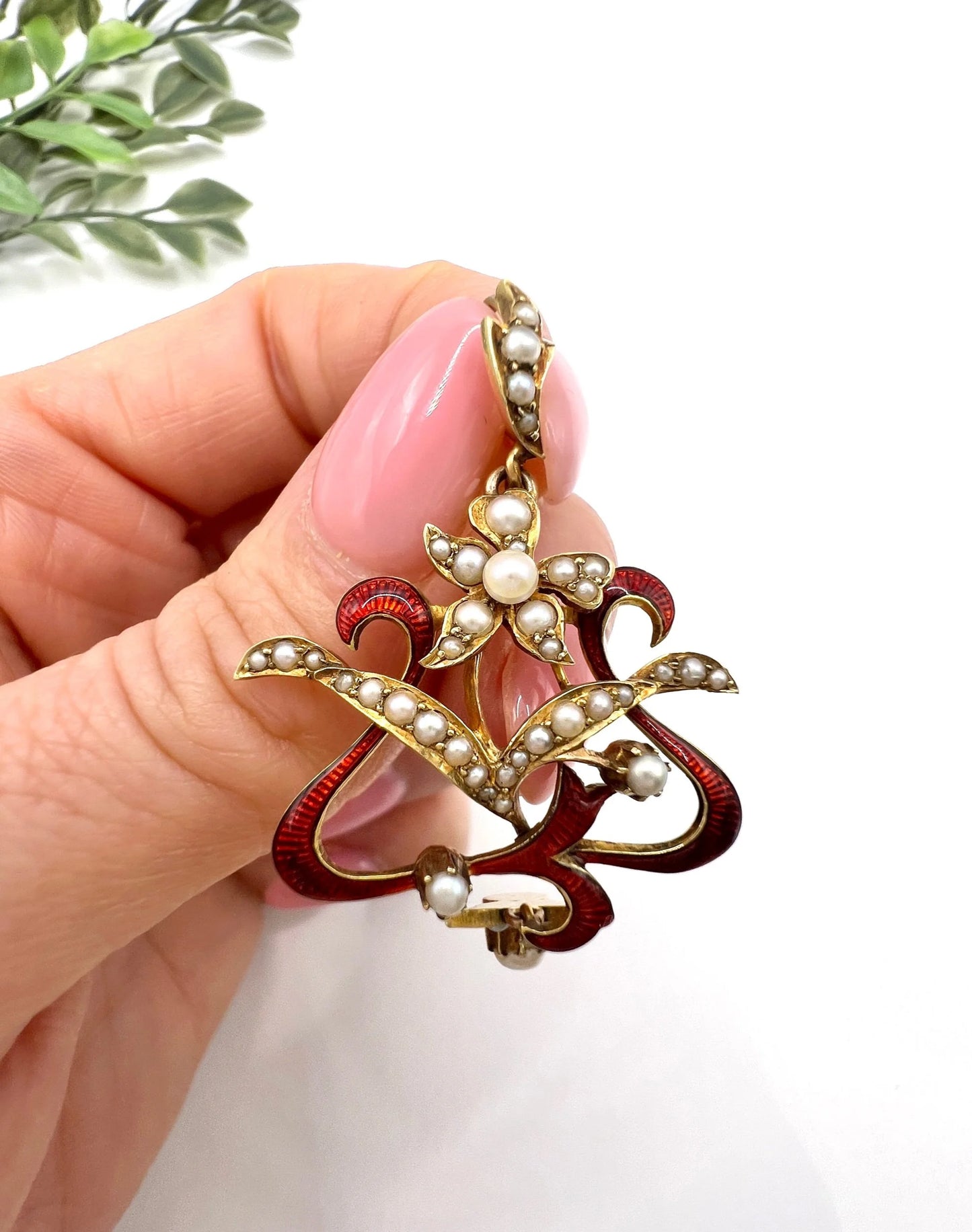 A floral pendant in yellow gold with seed pearls and red enamel, held in a hand