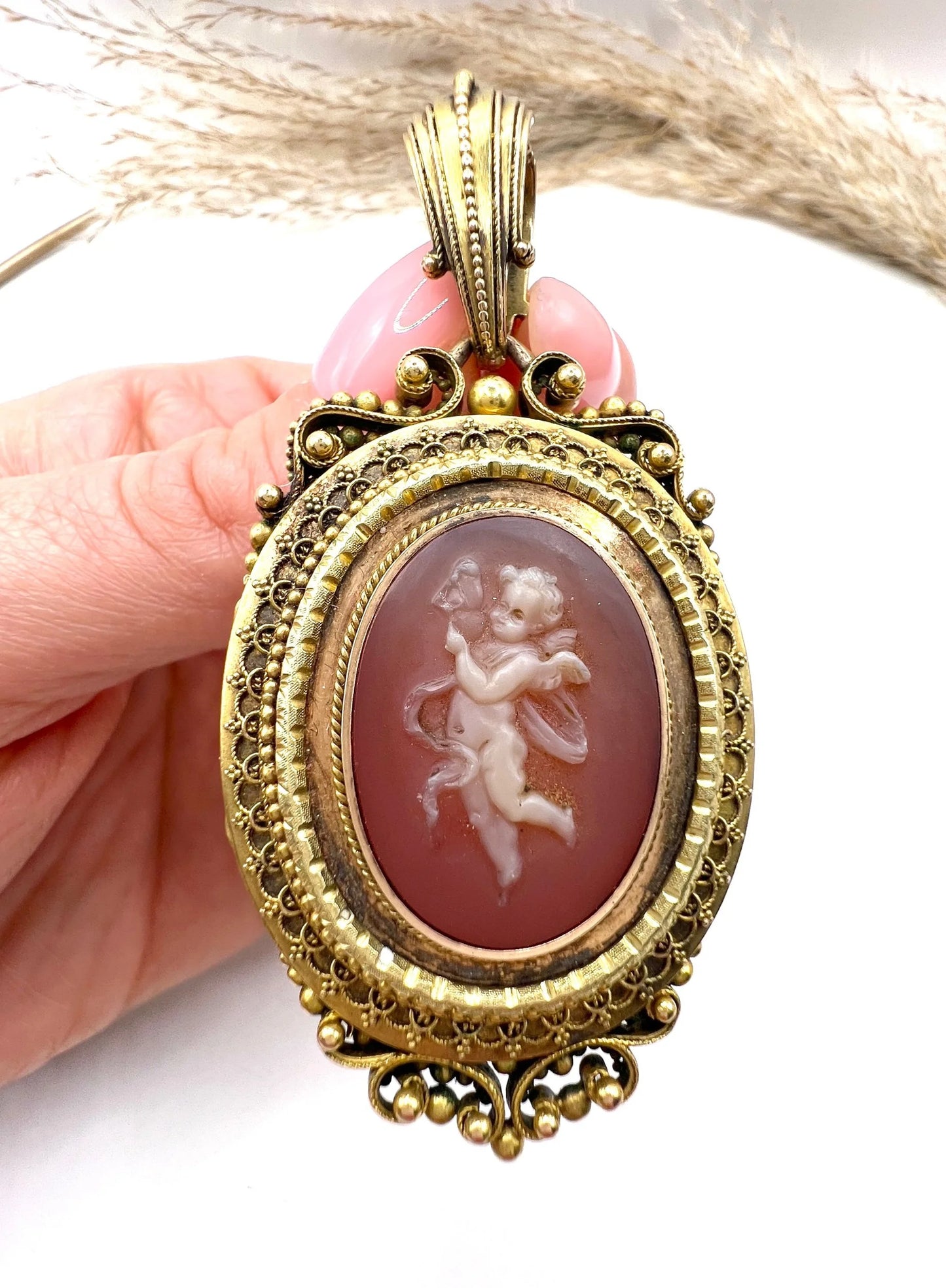 An ornate yellow gold oval cherub cameo locket pendant, held in a hand