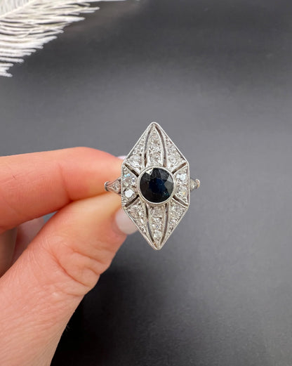 A navette shaped cluster cocktail ring with a sapphire centre surrounded by diamonds, in platinum, held in a hand