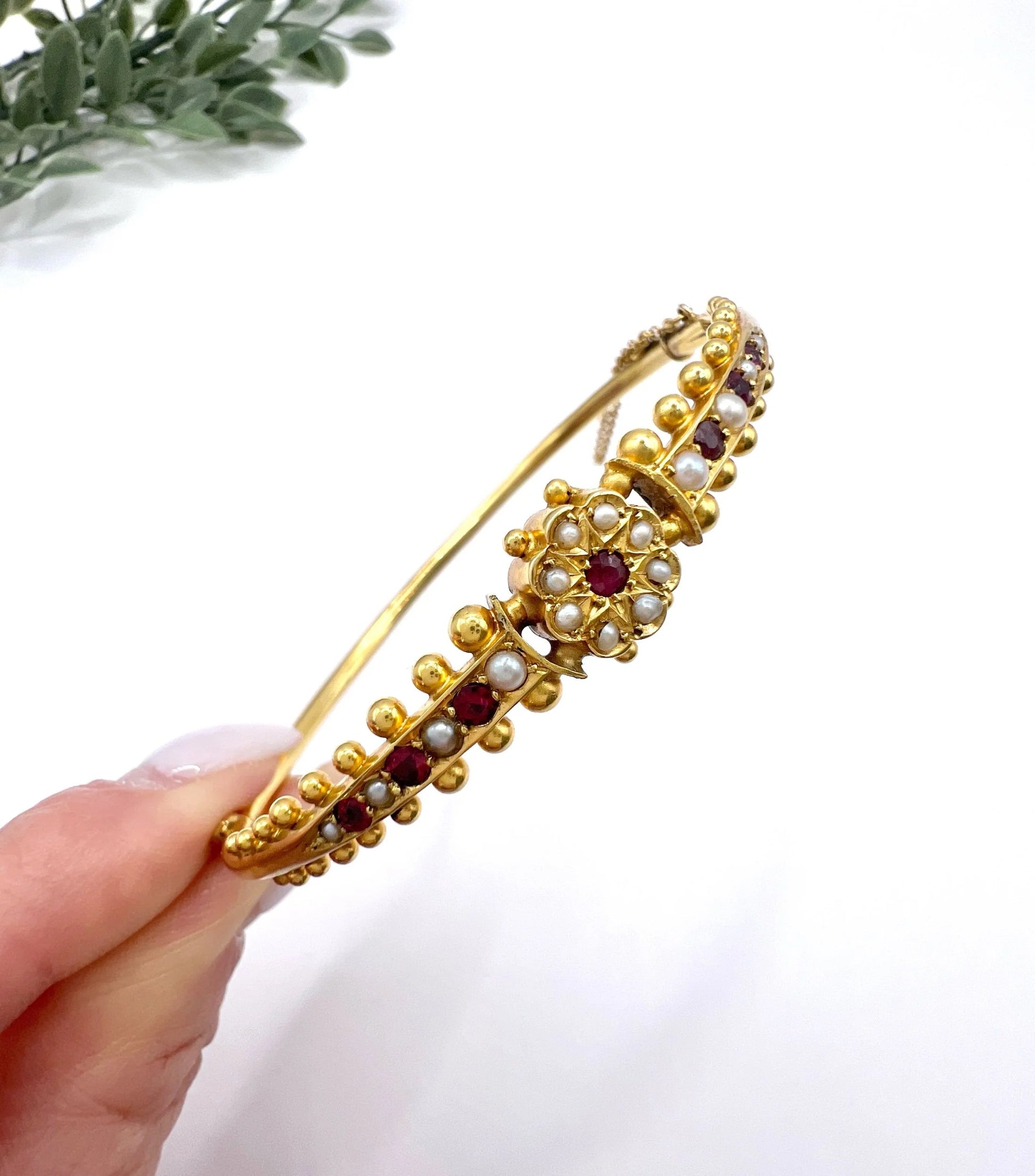 A yellow gold bangle bracelet set with rubies and seed pearls, held in a hand