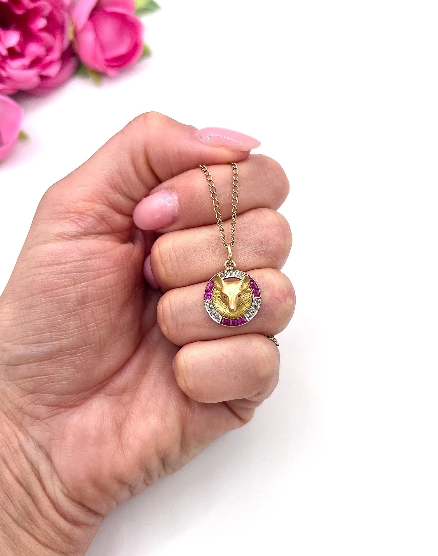 A fox head pendant in yellow gold with a ruby and diamond border, with a gold chain, held in a hand
