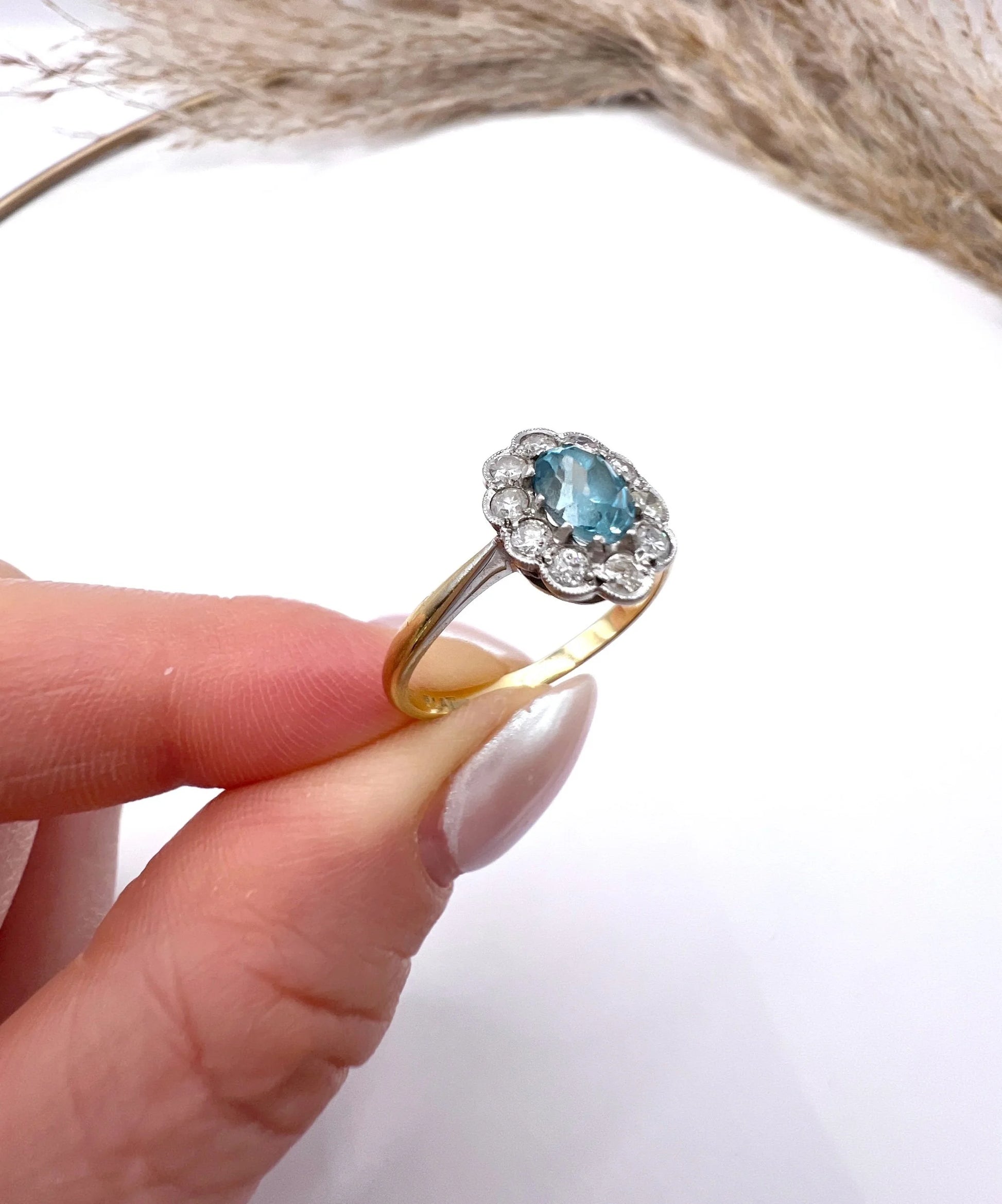 An oval cluster ring with a blue zircon centre surrounded by round diamonds, in yellow gold and platinum, held in a hand