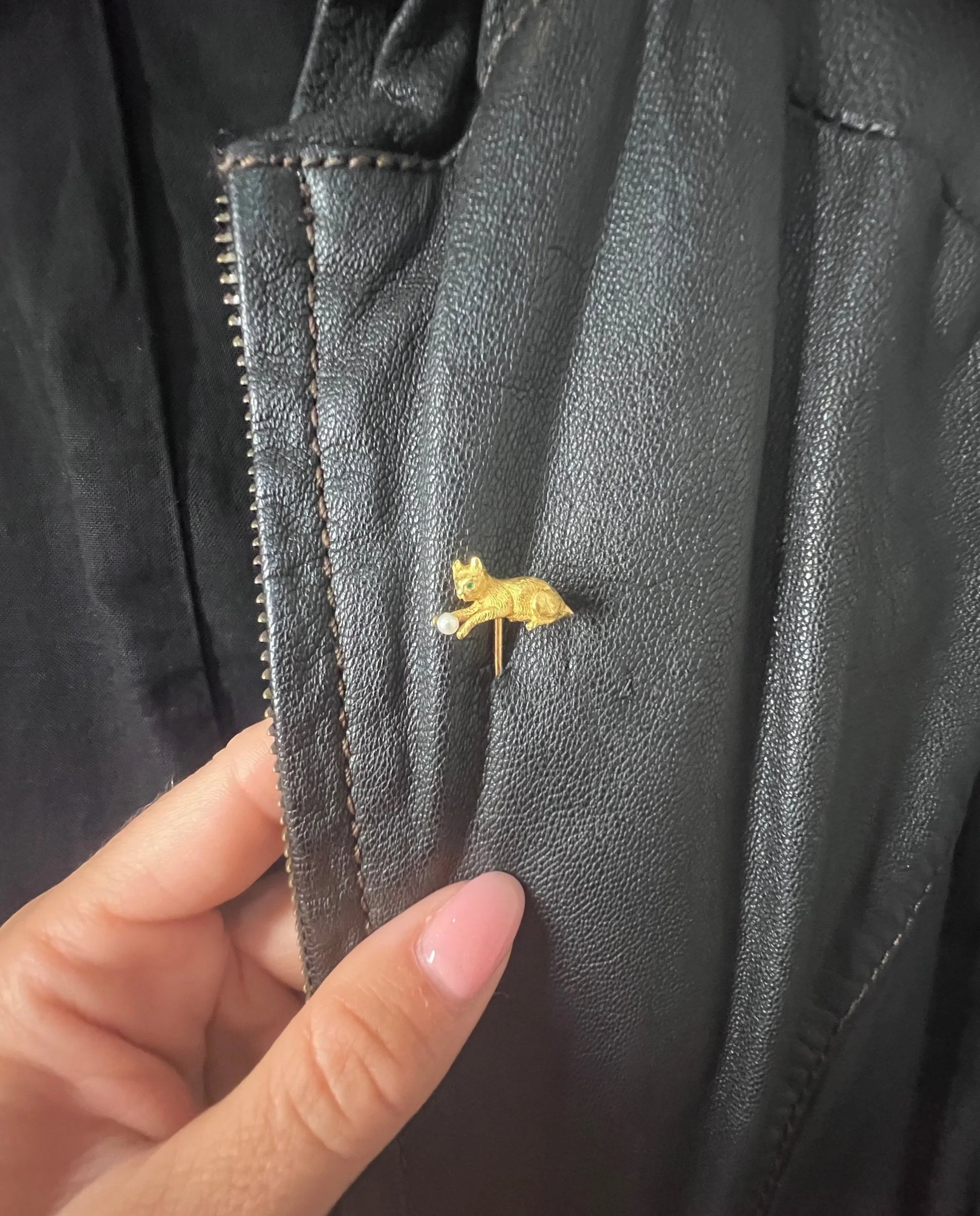 A yellow gold cat playing with a pearl ball stick pin, modelled on a black jacket