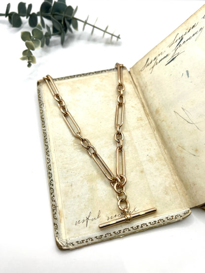 A rose gold trombone and fetter link Albert chain lying on top of an antique book, on a white background