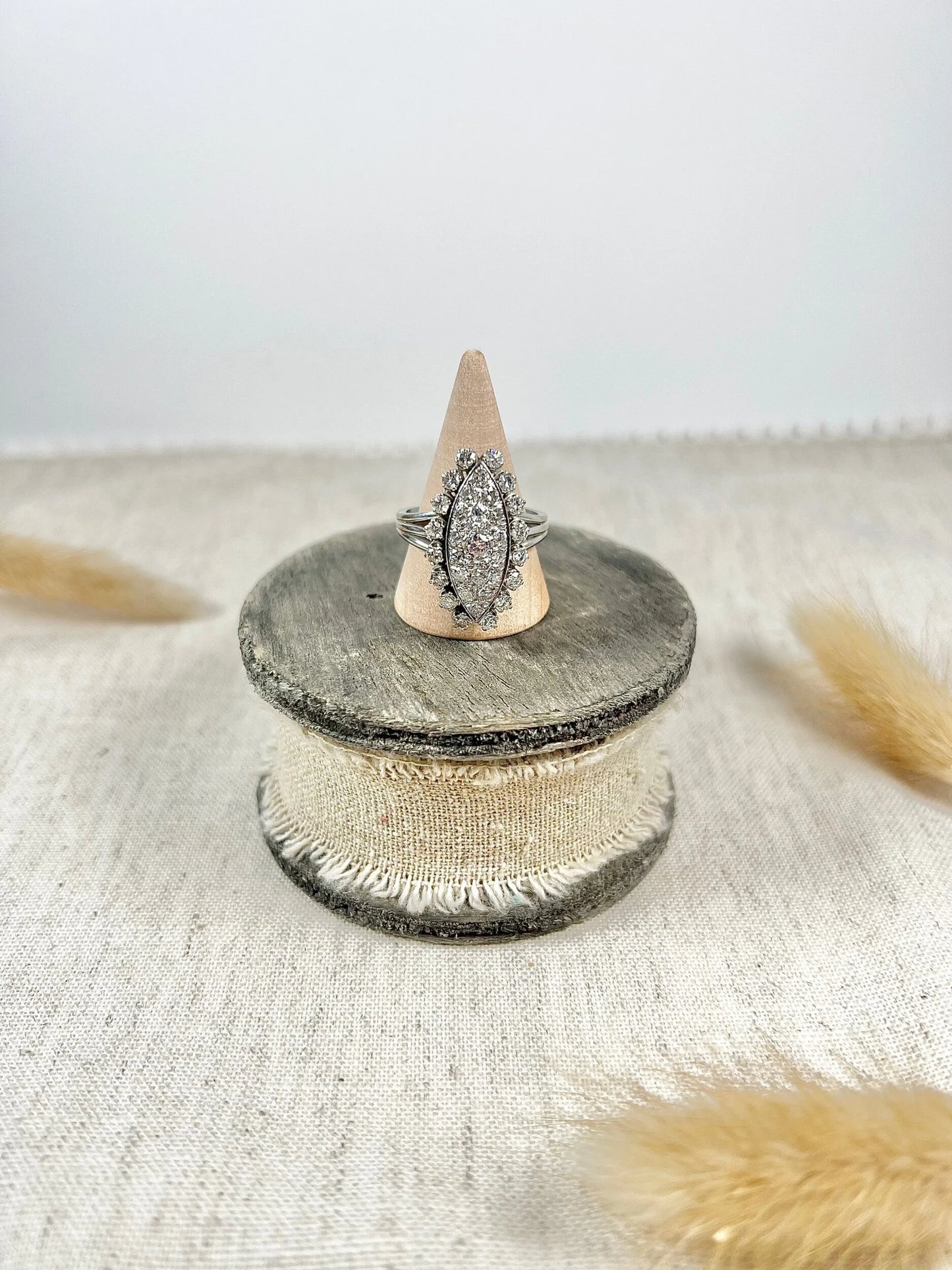 A navette shaped diamond cluster ring in white metal, on a ring cone, sitting on top of a wooden and cloth object