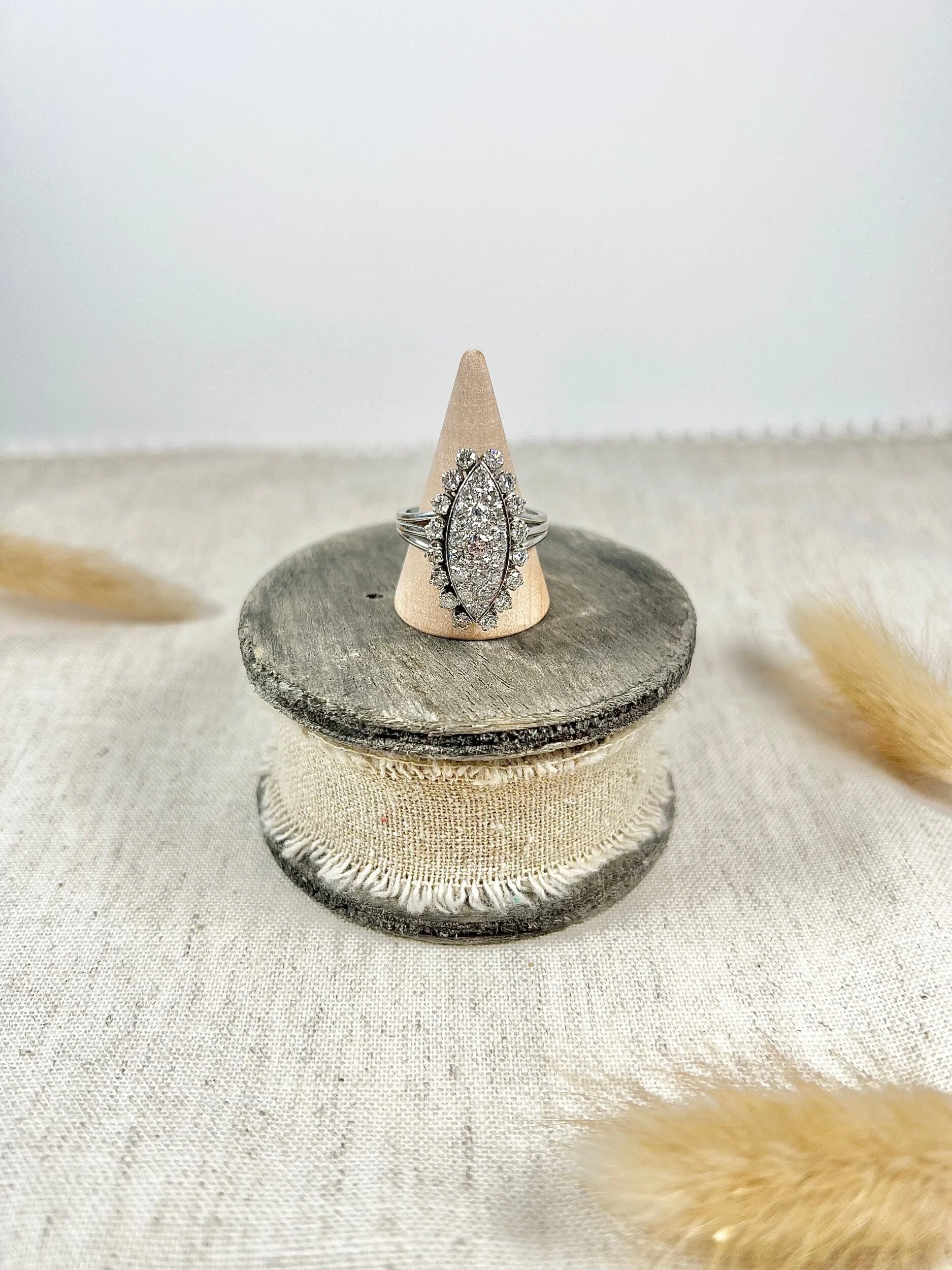 A navette shaped diamond cluster ring in white metal, on a ring cone, sitting on top of a wooden and cloth object