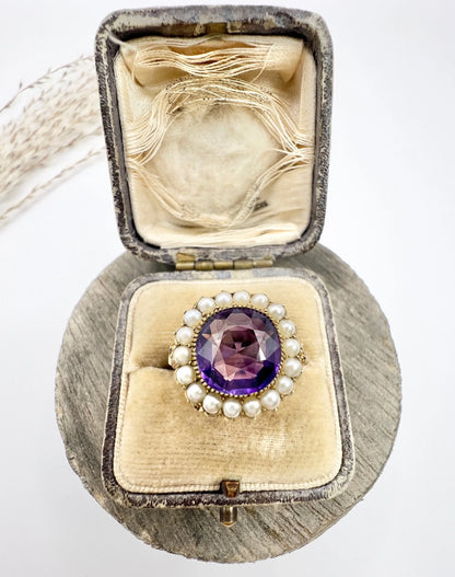 A cushion shaped amethyst cluster ring with a pearl border, in yellow gold, in a ring box