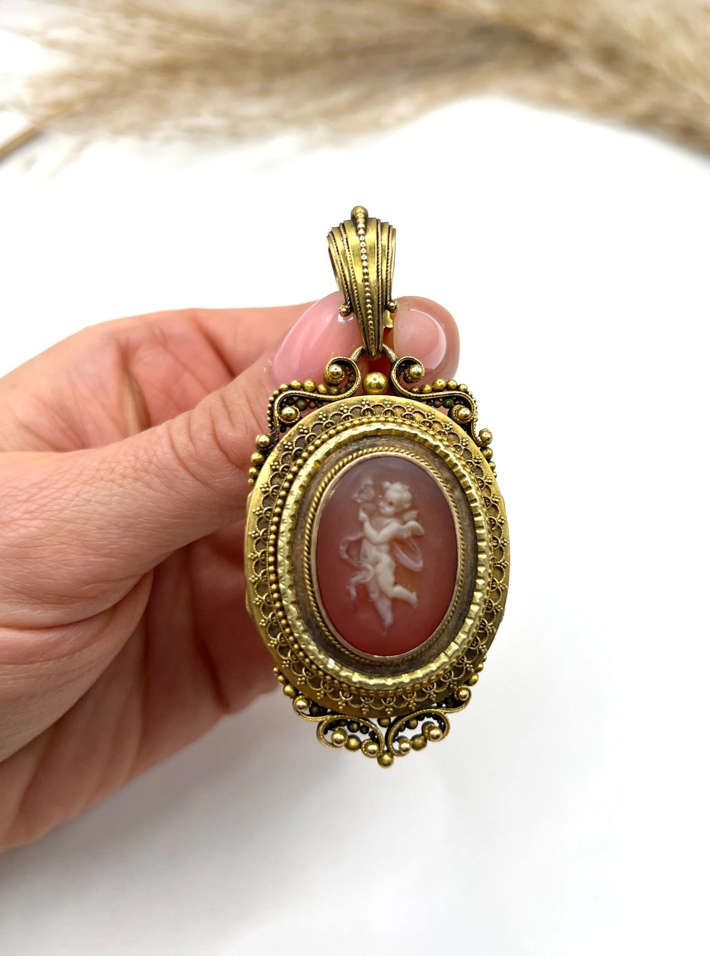 An ornate yellow gold oval cherub cameo locket pendant, held in a hand