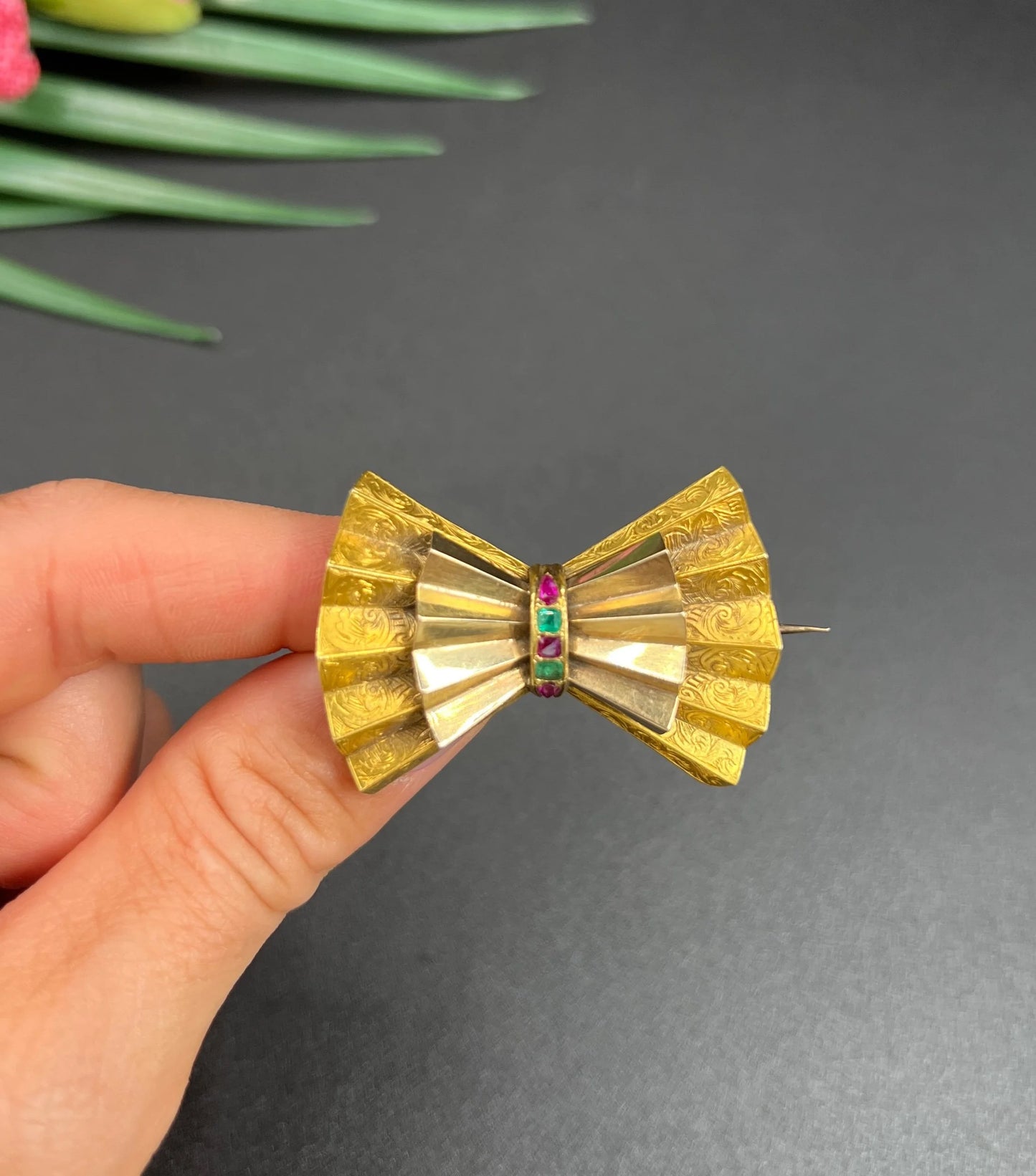 A yellow gold bow shaped brooch with rubies and emeralds, held in a hand