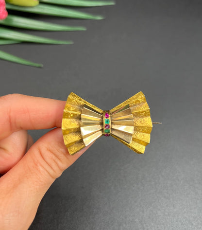 A yellow gold bow shaped brooch with rubies and emeralds, held in a hand