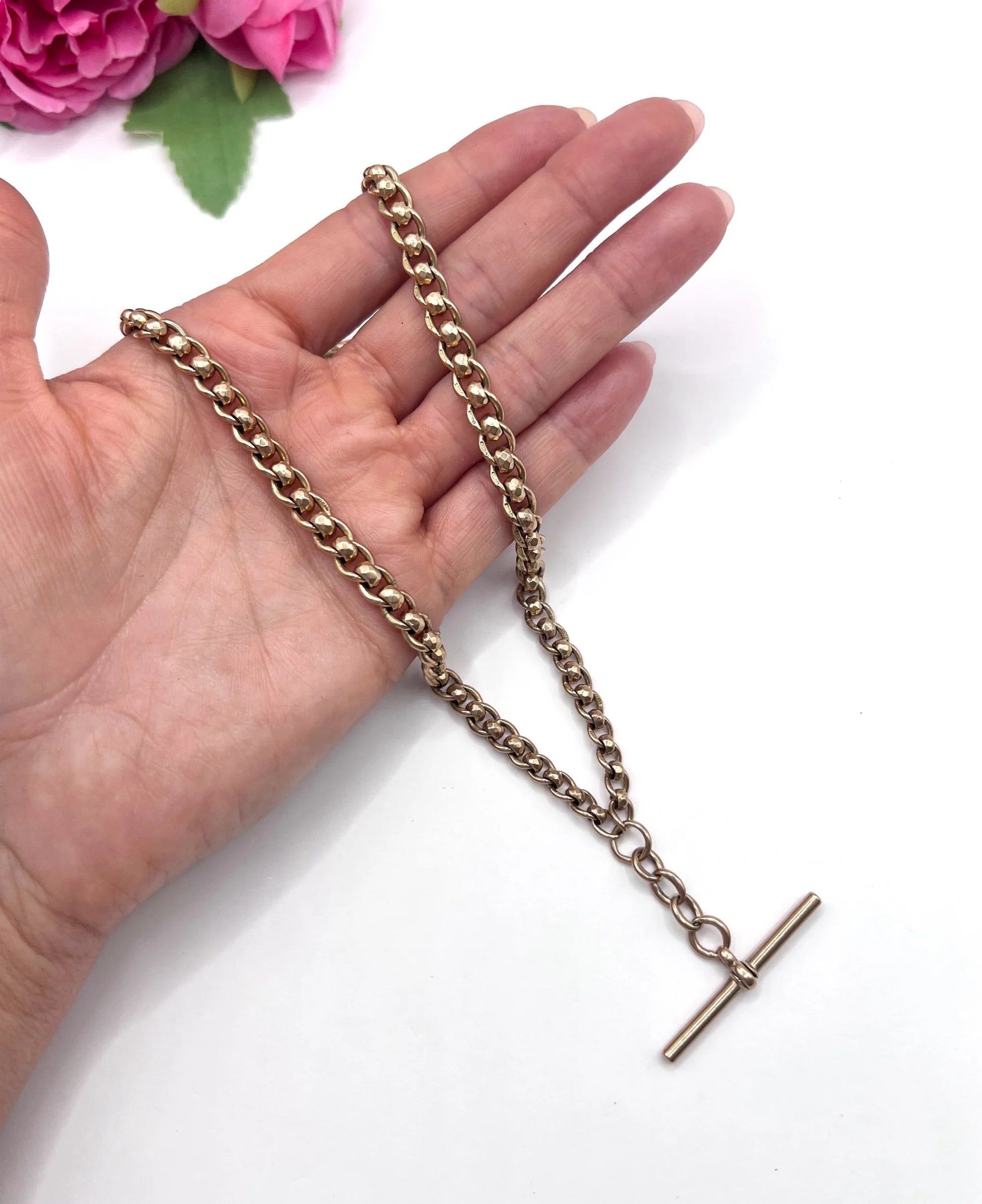 A rose gold fancy link Albert chain necklace with a T-bar attachment, held in a hand