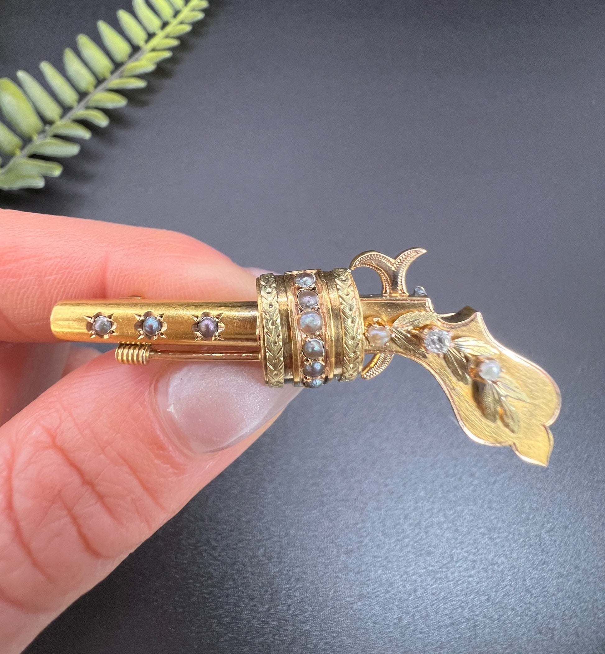 A yellow gold novelty gun brooch with diamonds and seed pearls, held in a hand