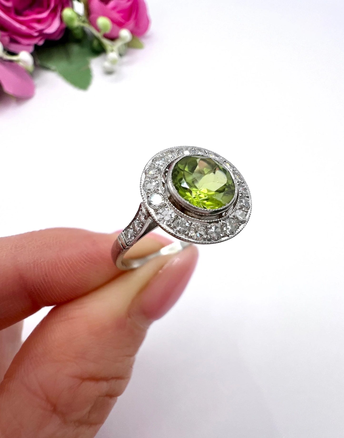 A white gold circular target cluster ring with a peridot centre surrounded by diamonds, held in a hand