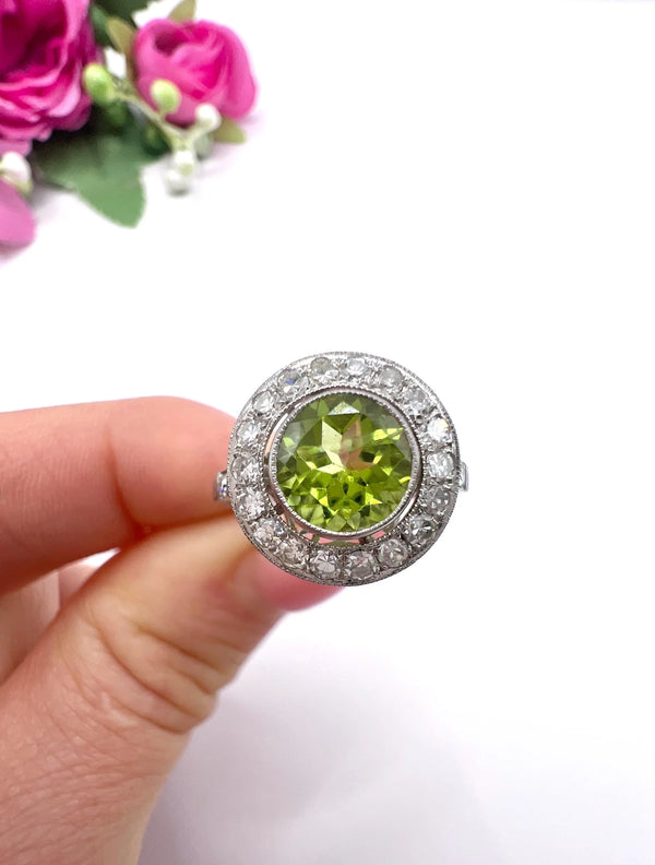 A white gold circular target cluster ring with a peridot centre surrounded by diamonds, held in a hand