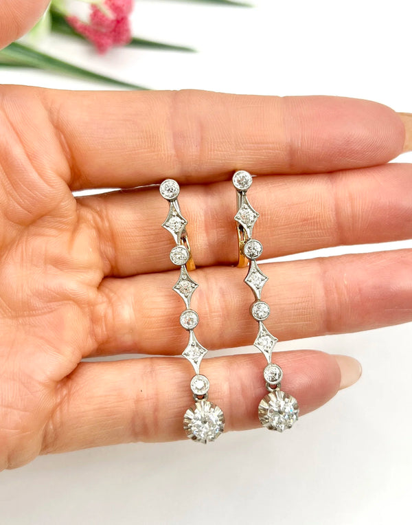 A pair of diamond drop earrings in yellow gold and platinum, held in a hand