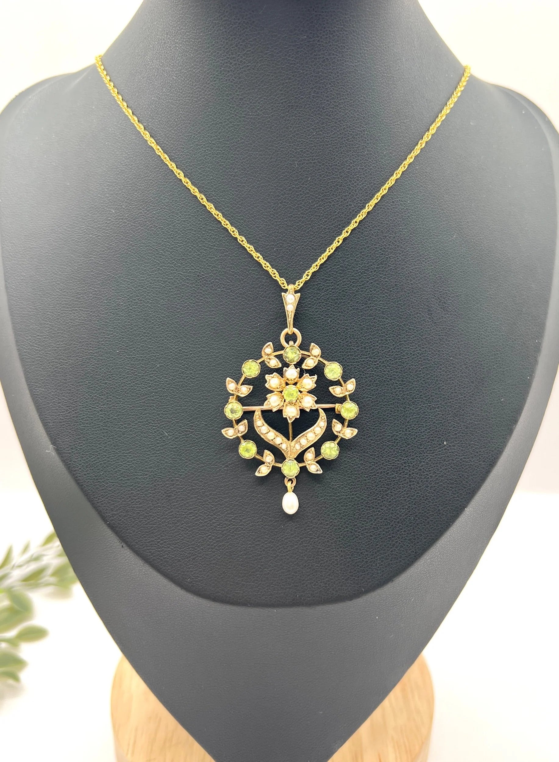 A peridot and pearl floral circle brooch/pendant in yellow gold, with a chain, on a black jewellery bust