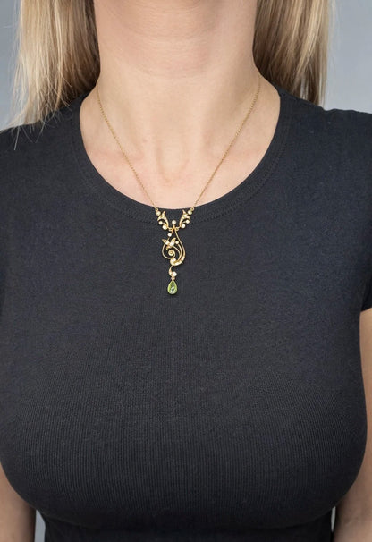 A yellow gold floral pendant necklace with pearls and a peridot drop, modelled by a woman wearing a black top