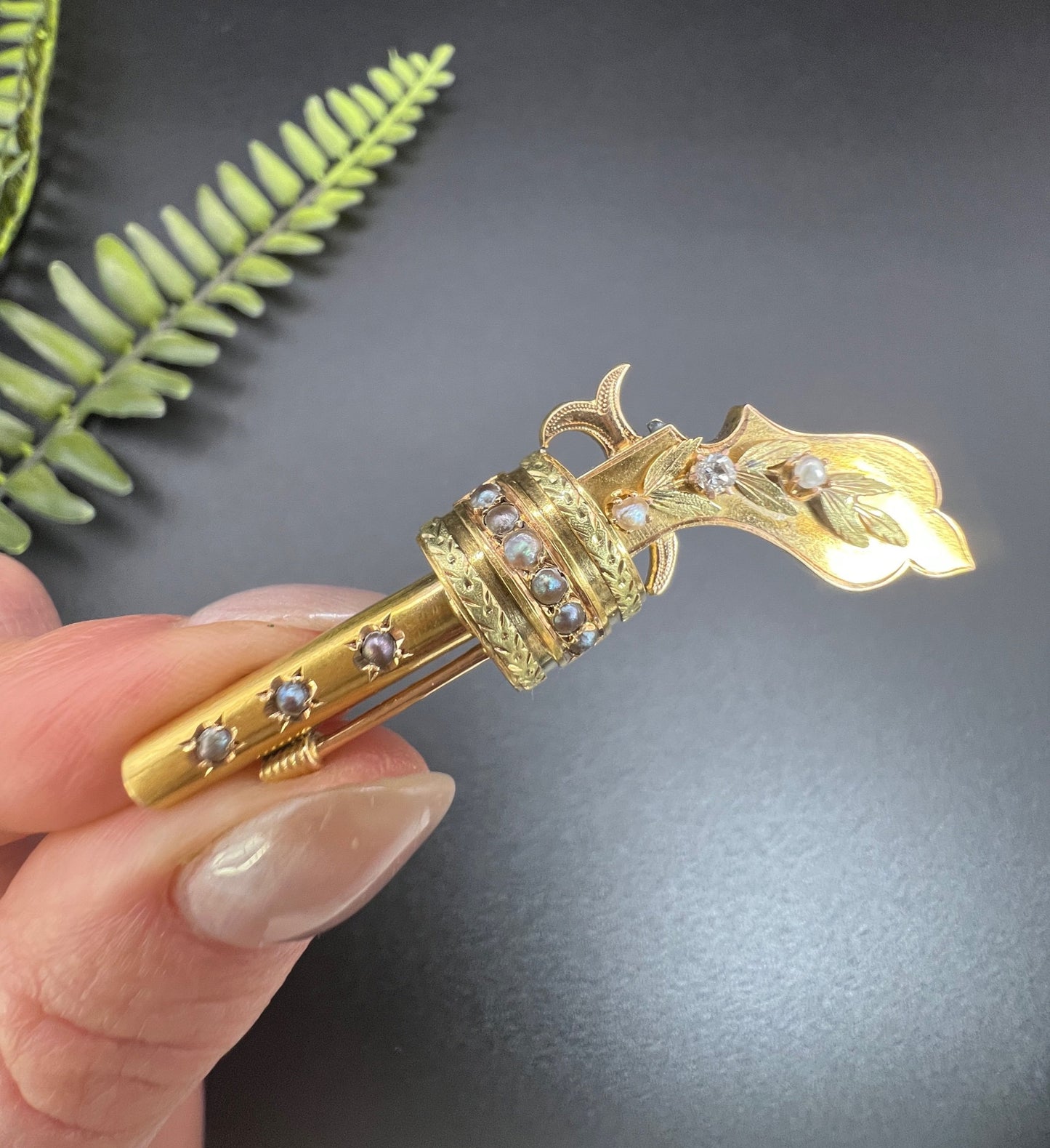 A yellow gold novelty gun brooch with diamonds and seed pearls, held in a hand