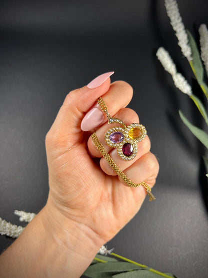 A yellow gold clover pendant with amethyst, citrine, garnet and seed pearls, with a chain, held in a hand