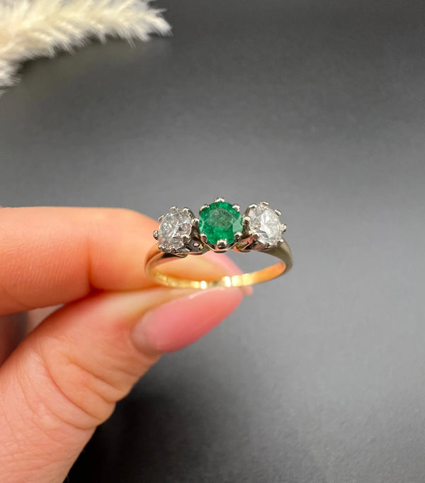 A three stone emerald and diamond ring in yellow gold and platinum, held in a hand