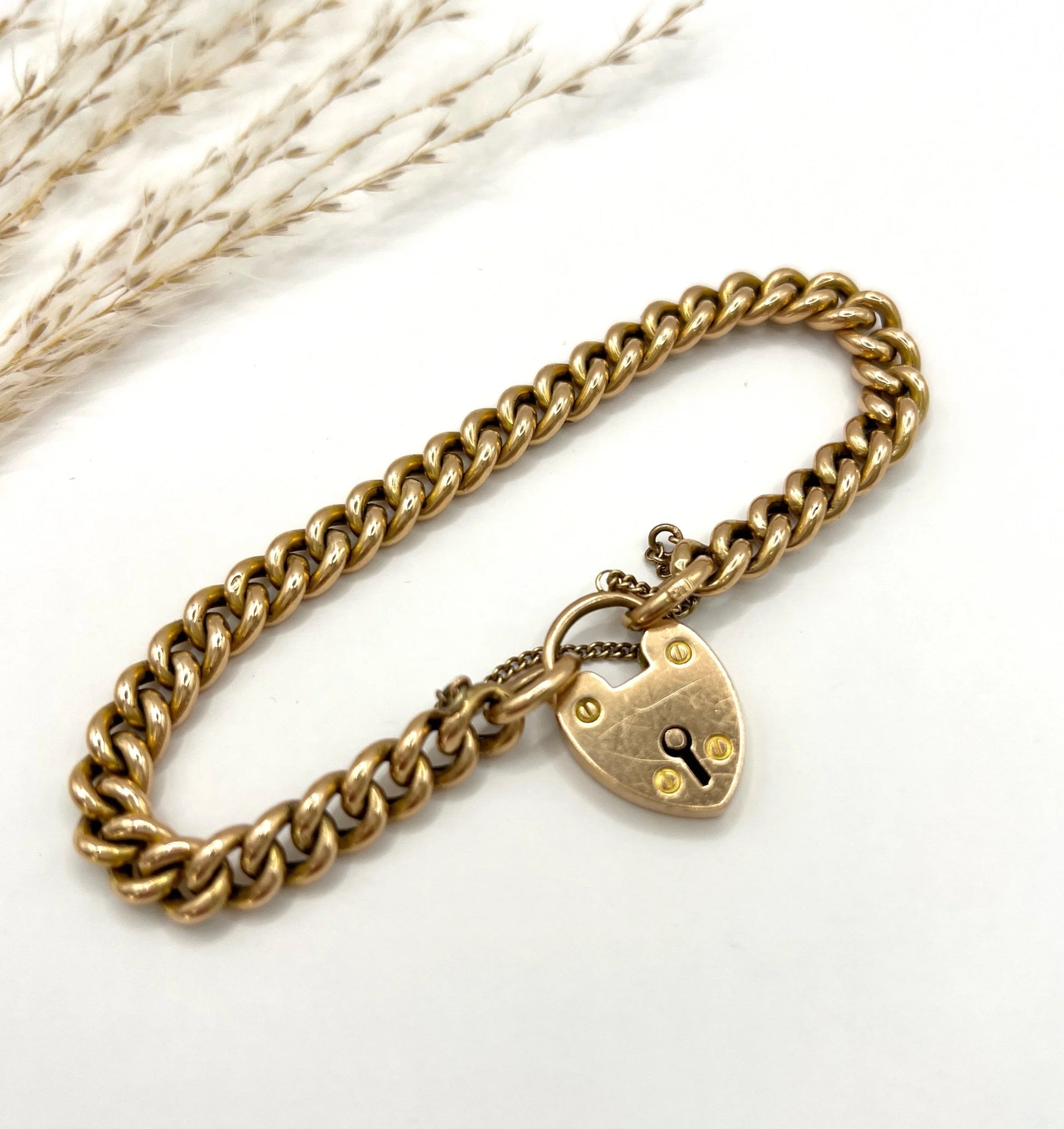 A gold curb link bracelet with heart shaped padlock, laid on a white background