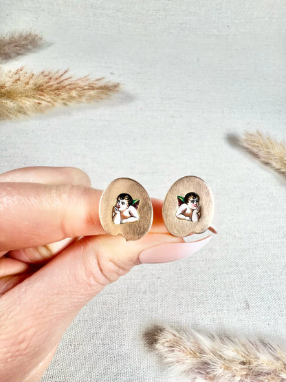 A pair of art palette shaped gold earrings with enamelled cherubs, held in a hand