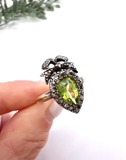 A pear cut peridot and round diamond cluster ring with bow detail, in yellow gold and white metal, in a hand