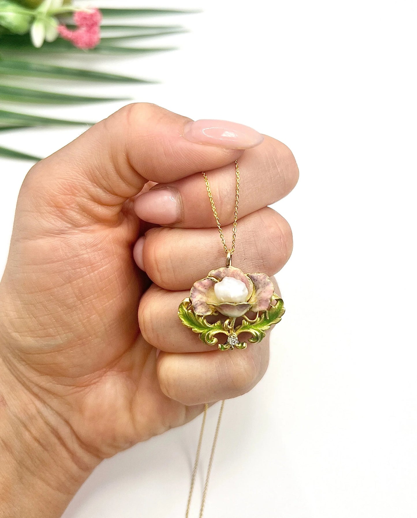 An enamel, diamond and pearl flower pendant with a gold chain, held in a hand