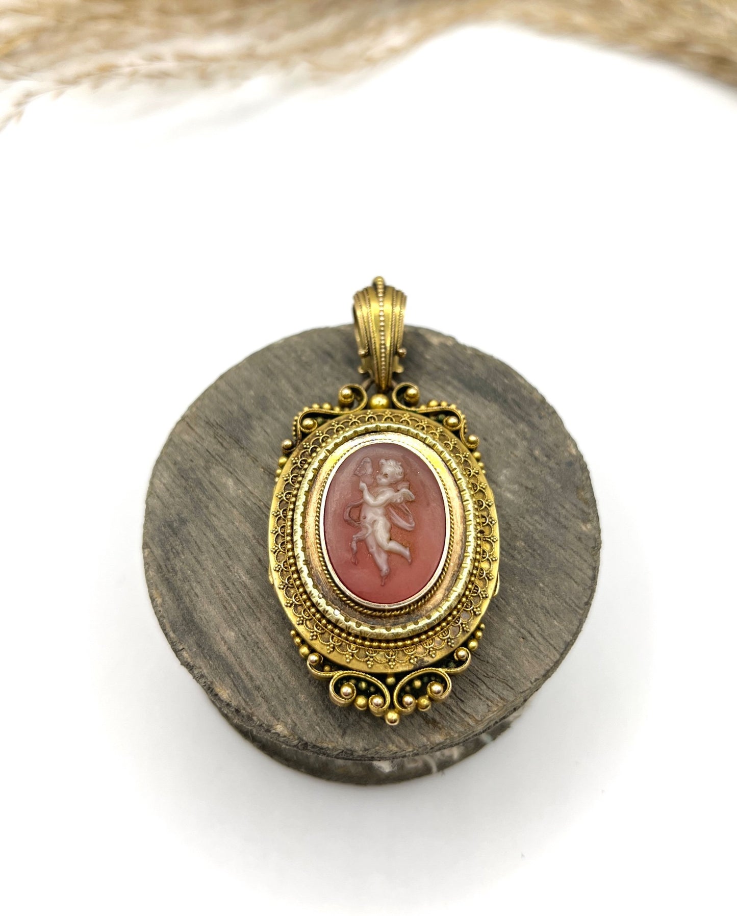 An ornate yellow gold oval cherub cameo locket pendant, on a wooden spool
