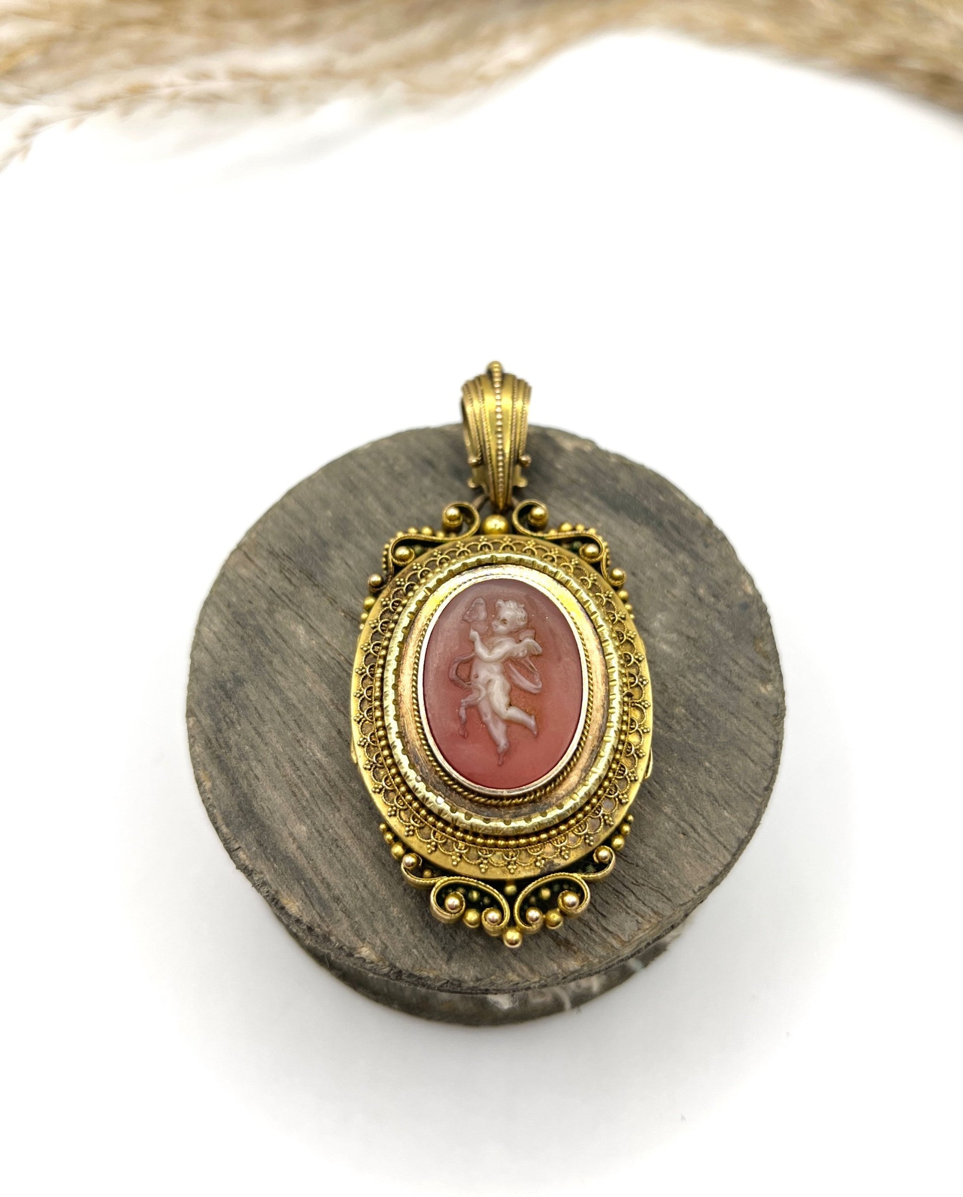 An ornate yellow gold oval cherub cameo locket pendant, on a wooden spool