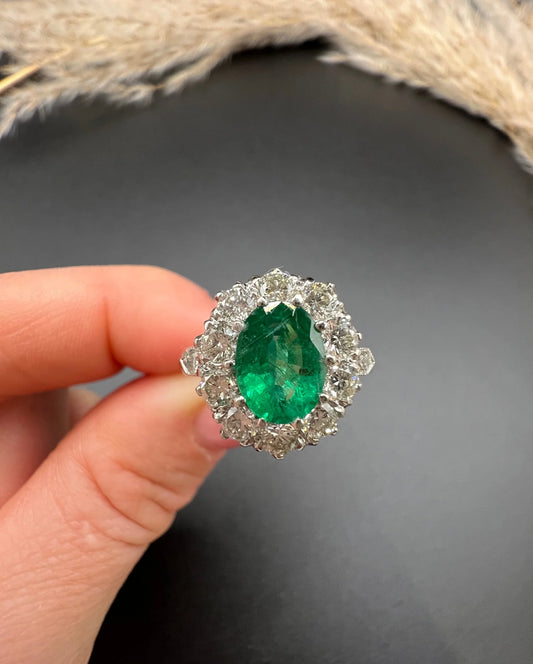 An oval emerald and diamond cluster ring in platinum, held in a hand