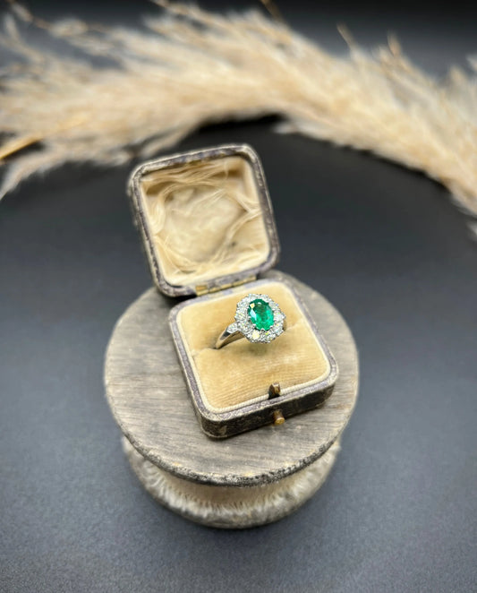 An oval emerald and diamond cluster ring in platinum, in an antique ring box