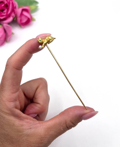 A yellow gold cat playing with a pearl ball stick pin, held in a hand