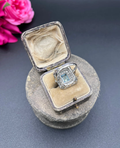 An octagonal shaped cluster ring with an aquamarine centre surrounded by two rows of diamonds, in platinum, in a ring box