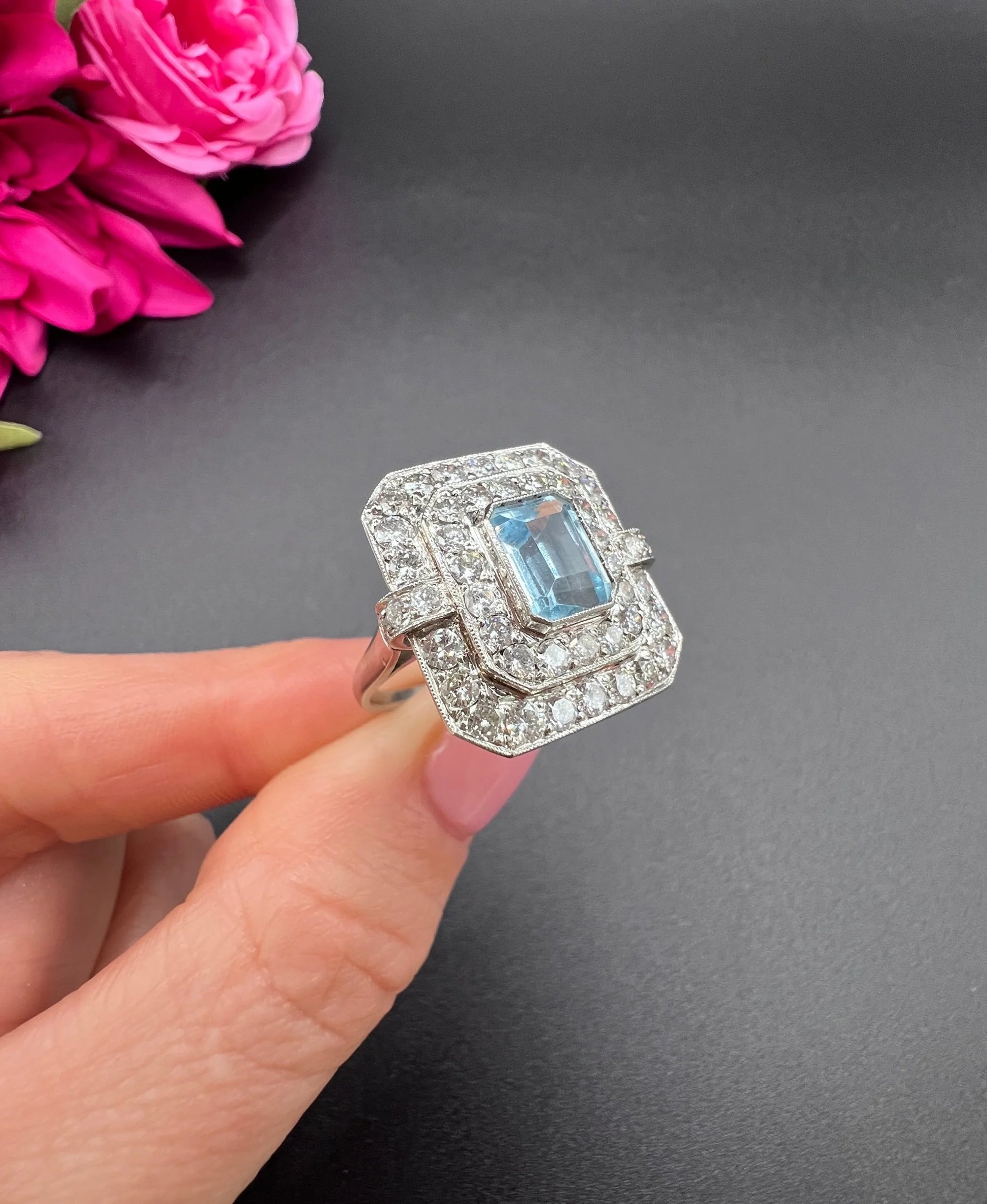 An octagonal shaped cluster ring with an aquamarine centre surrounded by two rows of diamonds, in platinum, held in a hand