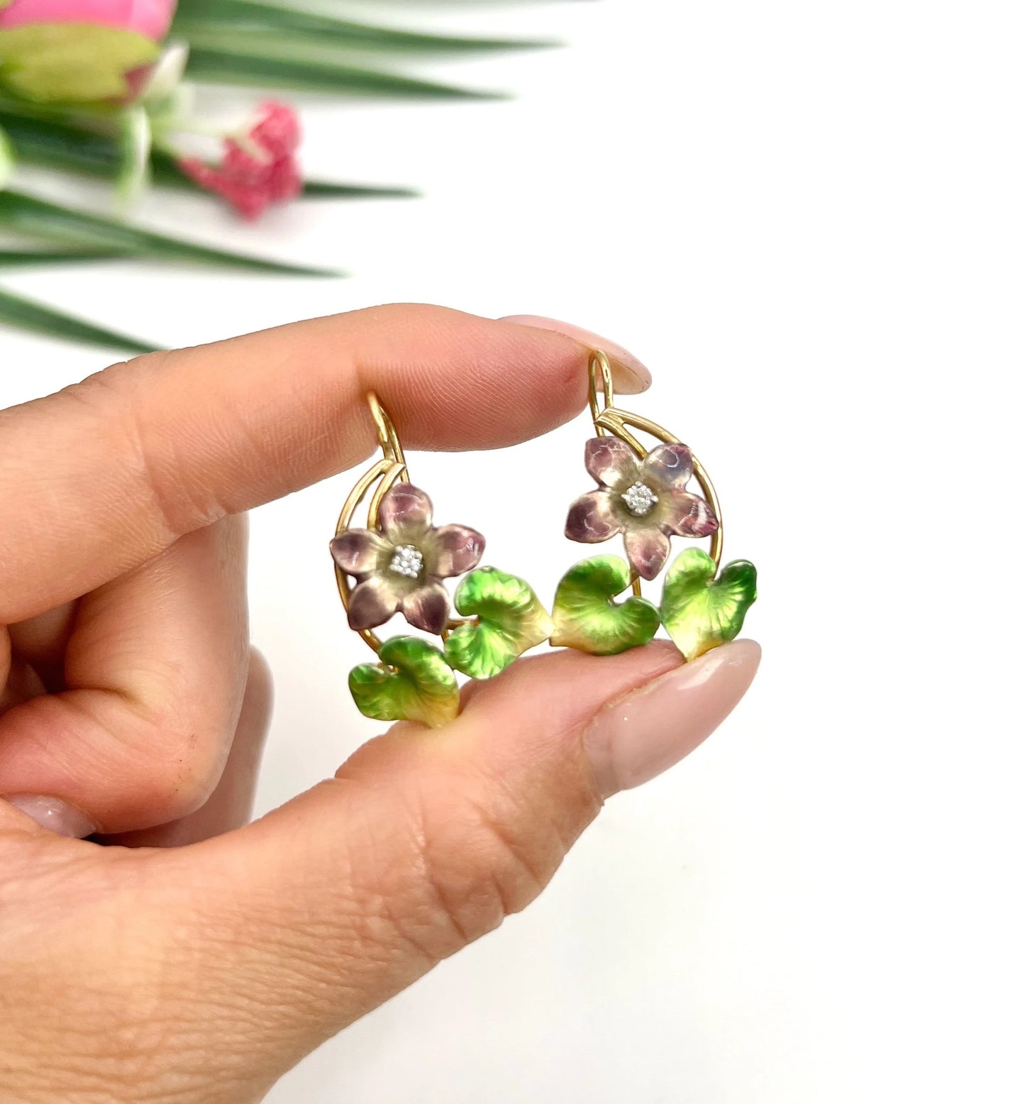 A pair of gold and enamel flower earrings with diamonds, held in a hand