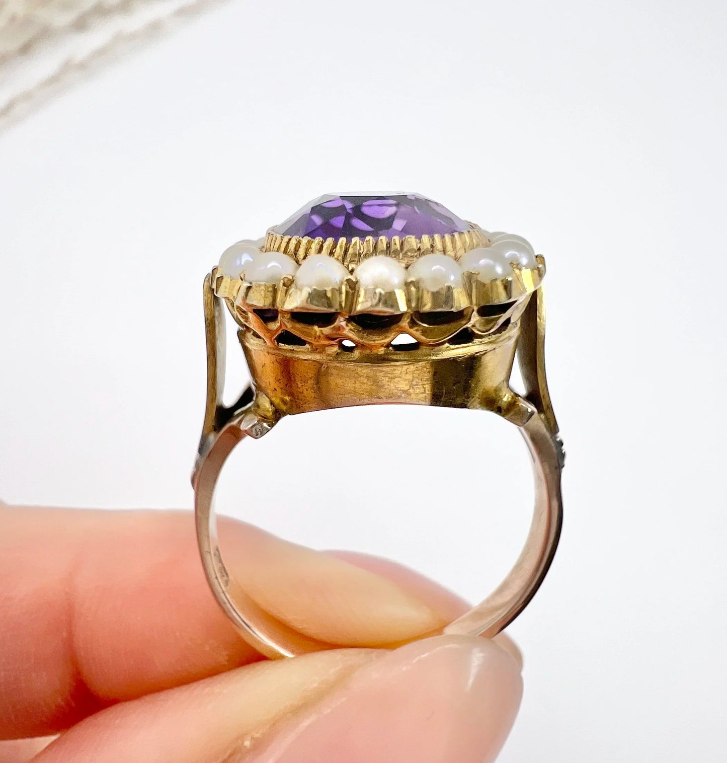 A cushion shaped amethyst cluster ring with a pearl border, in yellow gold, held in a hand