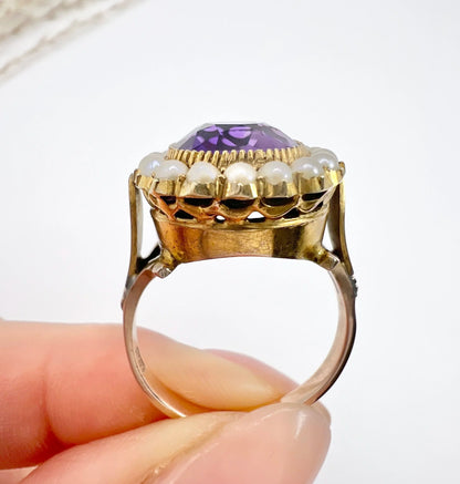 A cushion shaped amethyst cluster ring with a pearl border, in yellow gold, held in a hand