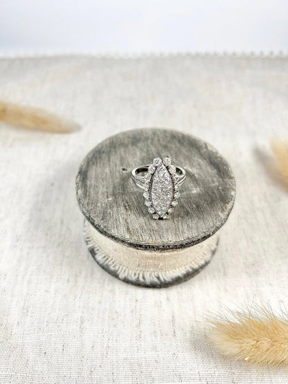 A navette shaped diamond cluster ring in white metal, sitting on top of a wooden and cloth object