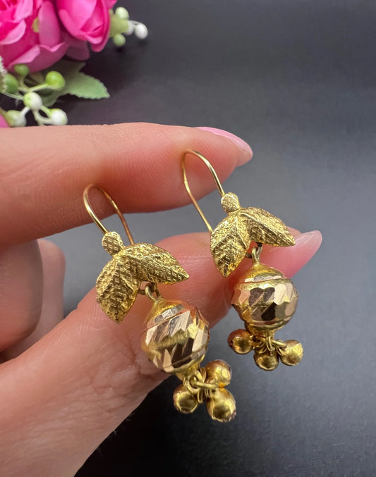 A pair of yellow gold leaf and faceted ball drop earrings with hook fittings, held in a hand