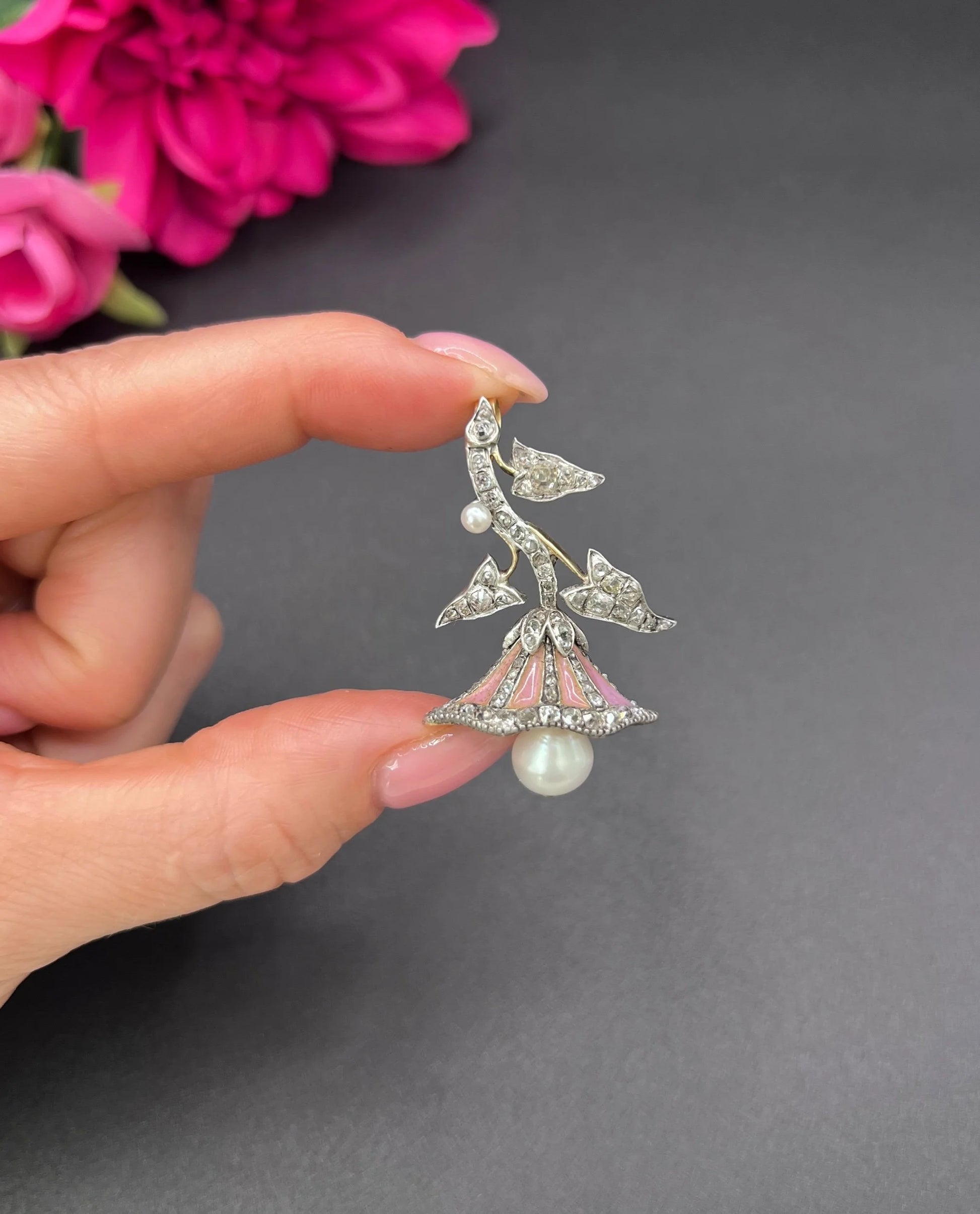 A pink enamel, diamond and pearl flower pendant in gold and silver held in a hand