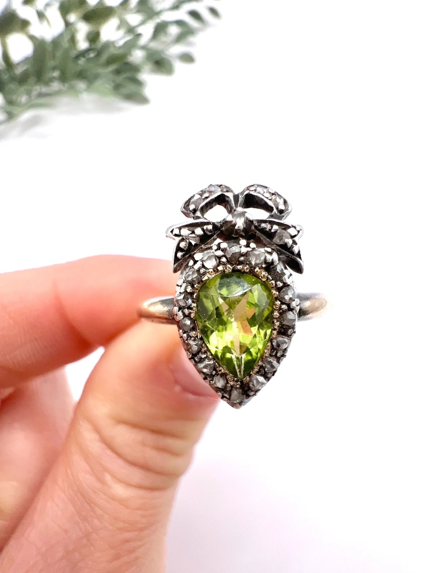 A pear cut peridot and round diamond cluster ring with bow detail, in yellow gold and white metal, held in a hand