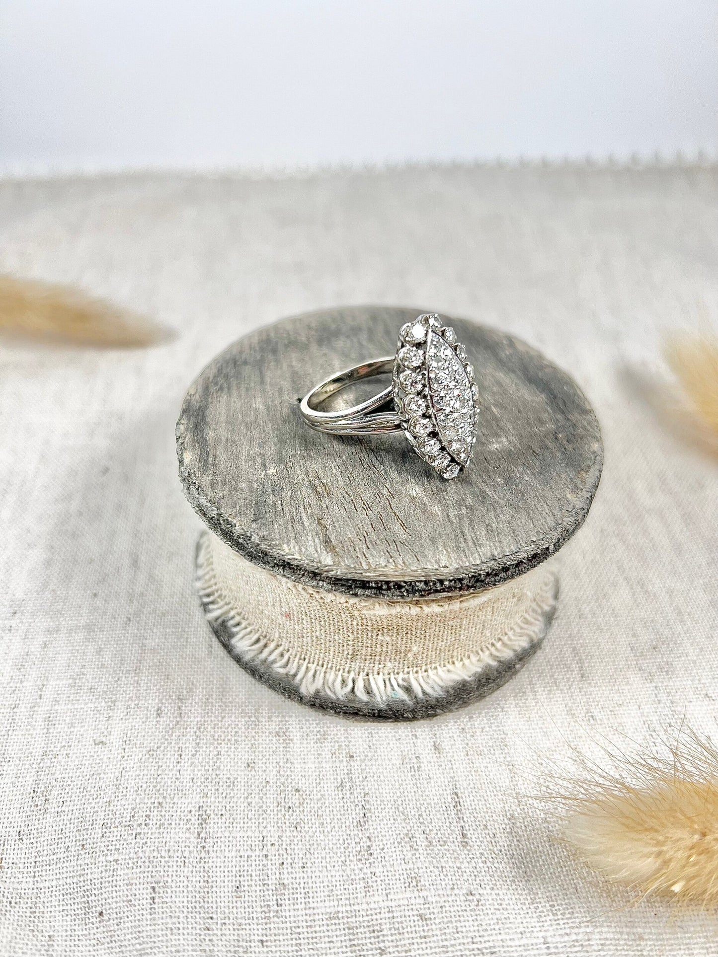 A navette shaped diamond cluster ring in white metal, sitting on top of a wooden and cloth object
