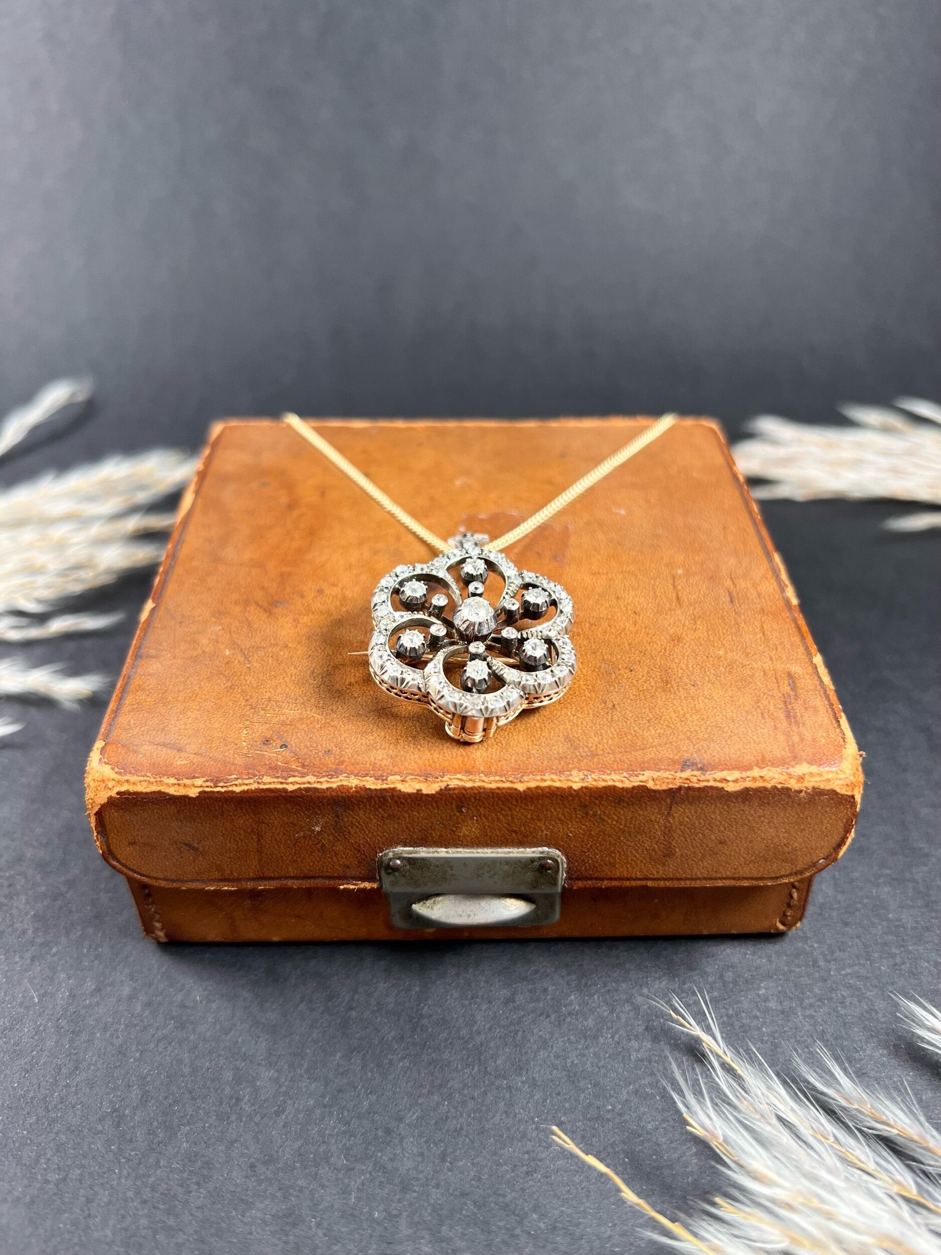 A floral design diamond set pendant/brooch on a gold chain on an antique brown jewellery box