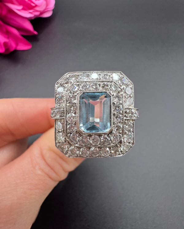 An octagonal shaped cluster ring with an aquamarine centre surrounded by two rows of diamonds, in platinum, held in a hand