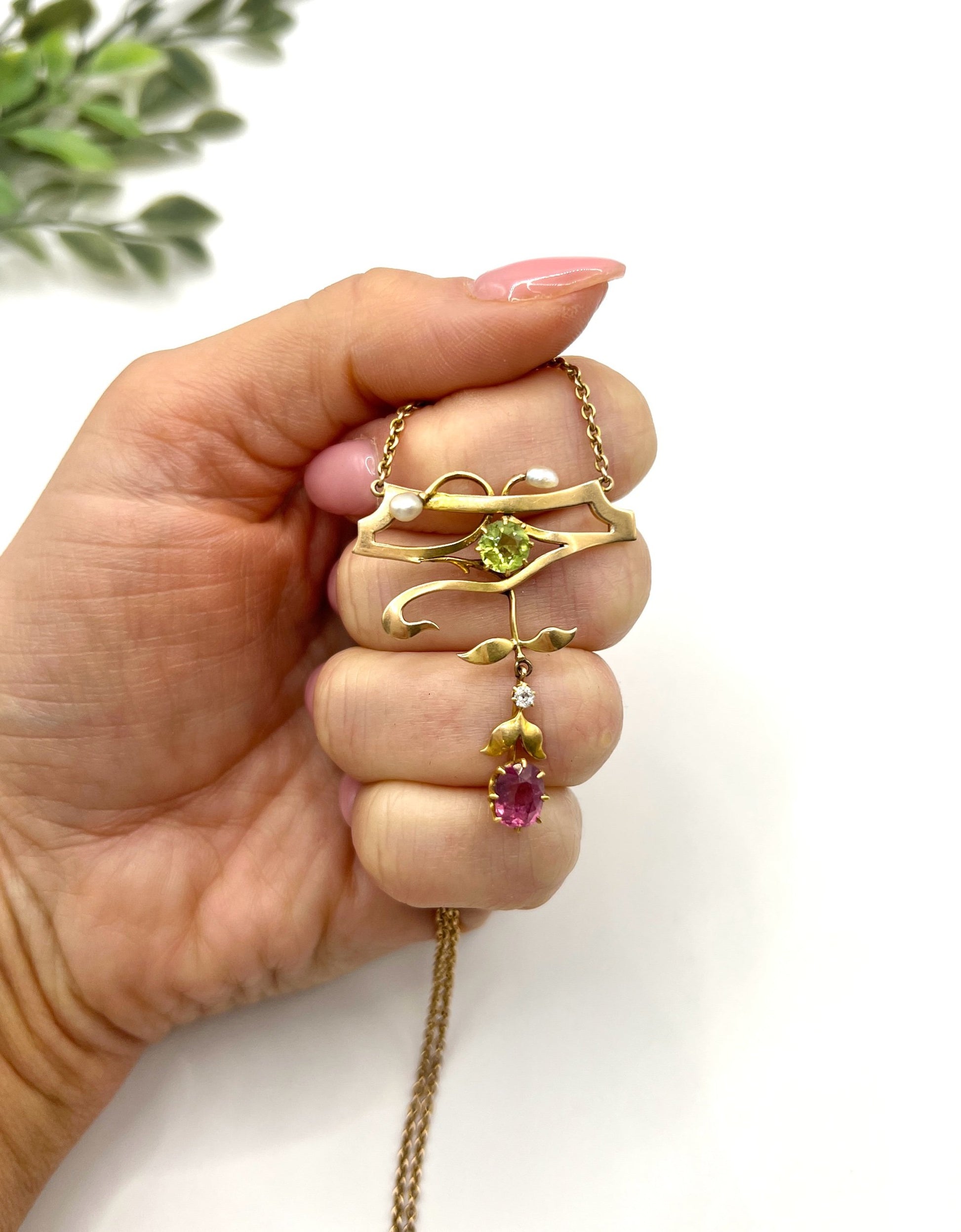 A yellow gold pendant necklace with pearls, a peridot, a diamond and a pink tourmaline drop, held in a hand